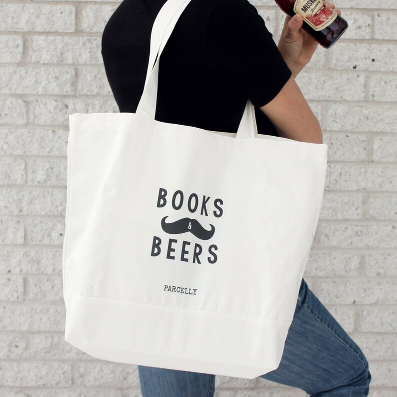 May include: A white canvas tote bag with black text that reads "BOOKS & BEERS" above a mustache graphic. The word "PARCELLY" is printed below. The bag has two long straps and is being held by a person.