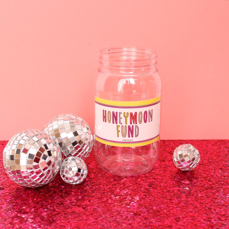 May include: A clear glass jar with a label that reads "Honeymoon Fund" on a pink sparkly surface. The jar is surrounded by silver disco balls.