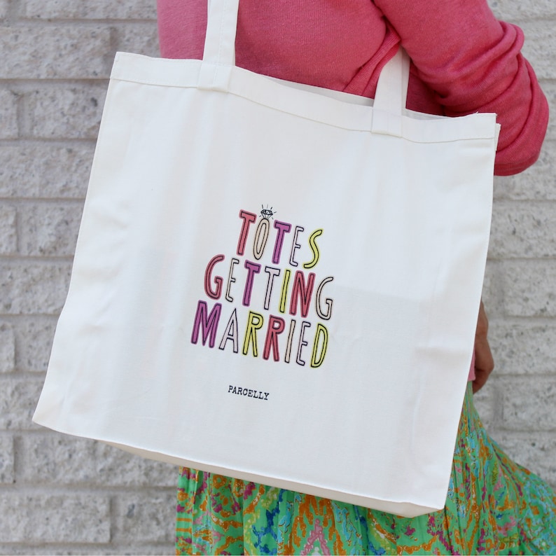 May include: White canvas tote bag with the text "Totes Getting Married" in colorful letters. The text is in a stacked, playful font. The bag has a black handle and the brand name "Parcelly" is printed in black at the bottom.