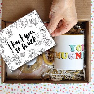 May include: A gift box filled with a mug that says "You Mugnificent", a cookie, and a card that says "Thank you so much!" The box is lined with gold and white shredded paper.