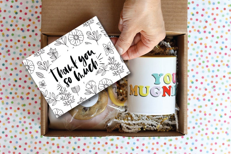 May include: A gift box filled with a mug that says "You Mugnificent", a cookie, and a card that says "Thank you so much!" The box is lined with gold and white shredded paper.