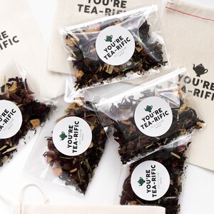 May include: Five small clear bags filled with loose leaf tea blends. Each bag has a round white sticker with the text "You're Tea-Rific" and a small green teapot icon. The bags are on a white surface.