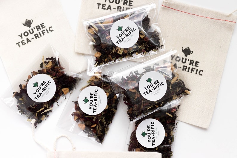 May include: Five small clear bags filled with loose leaf tea blends. Each bag has a round white sticker with the text "You're Tea-Rific" and a small green teapot icon. The bags are on a white surface.