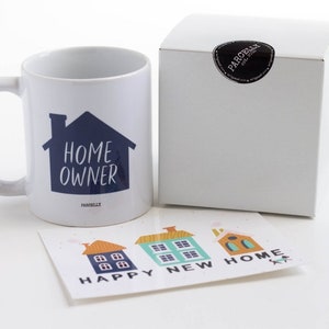May include: White ceramic mug with a blue house graphic and the text "HOME OWNER". The mug is packaged in a white box with a black sticker. A card with three colorful houses and the text "HAPPY NEW HOME" is also included.