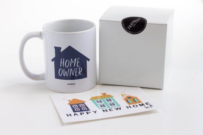 May include: White ceramic mug with a blue house graphic and the text "HOME OWNER". The mug is packaged in a white box with a black sticker. A card with three colorful houses and the text "HAPPY NEW HOME" is also included.