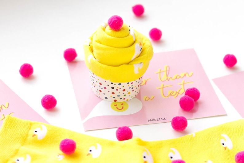 May include: A yellow cupcake-shaped sock gift with a pink pom-pom on top. The cupcake is sitting on a pink card with the text "Sweeter than a text" and the word "PARCELLY".