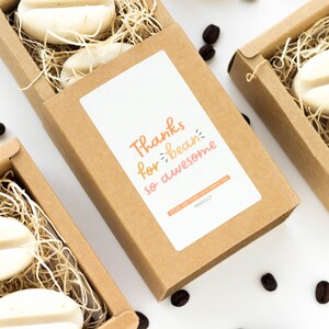 May include: Kraft paper boxes filled with straw and coffee bean-shaped soaps. The boxes have a white label with the text "Thanks for bean so awesome" in orange and pink. Scattered coffee beans are visible.