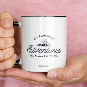 May include: White ceramic mug with black trim and handle, featuring the text "MY FAVORITE Adventures ARE ALWAYS WITH YOU" in black script. The mug has a mountain and sunburst graphic. The brand name "PARCELLY" is printed at the bottom.