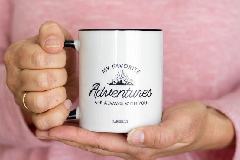 May include: White ceramic mug with black trim and handle, featuring the text "MY FAVORITE Adventures ARE ALWAYS WITH YOU" in black script. The mug has a mountain and sunburst graphic. The brand name "PARCELLY" is printed at the bottom.