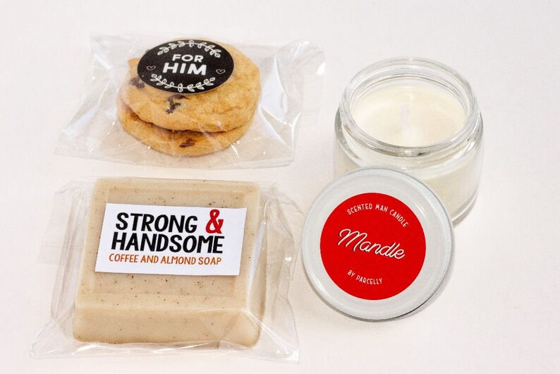 May include: A gift set featuring two chocolate chip cookies with a "For Him" sticker, a bar of coffee and almond soap, and a scented candle in a glass jar. The candle has a red label that says "Mandle".