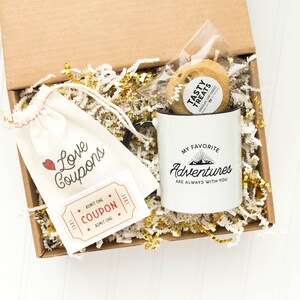 May include: A brown cardboard box filled with gold and white shredded paper. The box contains a white mug with black text that reads "My favorite adventures are always with you", a small white bag with the text "Love Coupons" and a red heart, a small white coupon that reads "Admit One Coupon Admit One", and a clear plastic bag with a cookie and a sticker that reads "Tasty Treats".