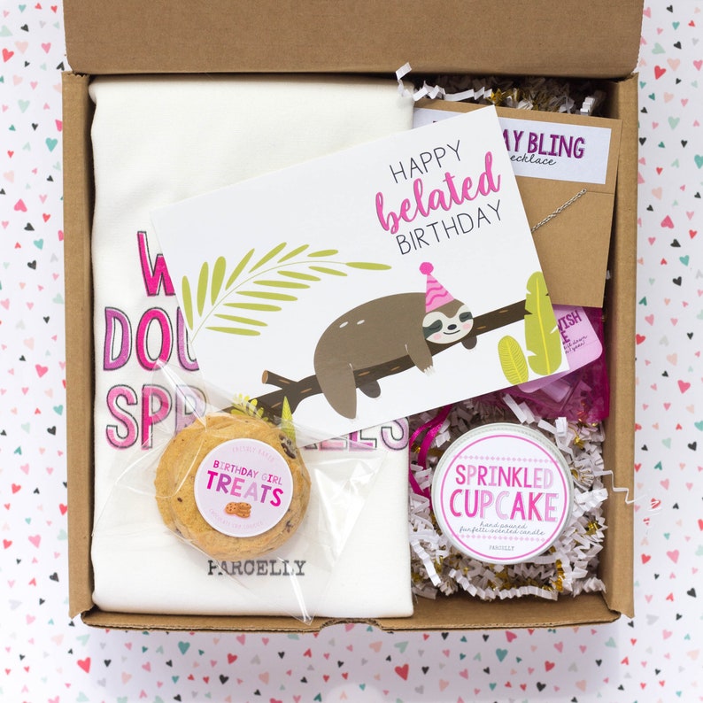 May include: A gift box containing a white t-shirt with pink text, a greeting card with a sloth illustration, a cookie, a candle, and a necklace. The card reads "Happy belated Birthday". The candle is labeled "Sprinkled Cupcake".