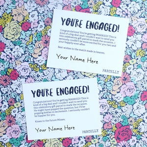 May include: Two white cards with a floral background. The cards have the text "You're Engaged!" and a message congratulating the couple on their engagement. The cards also have the text "Your Name Here" and "Parcellly".