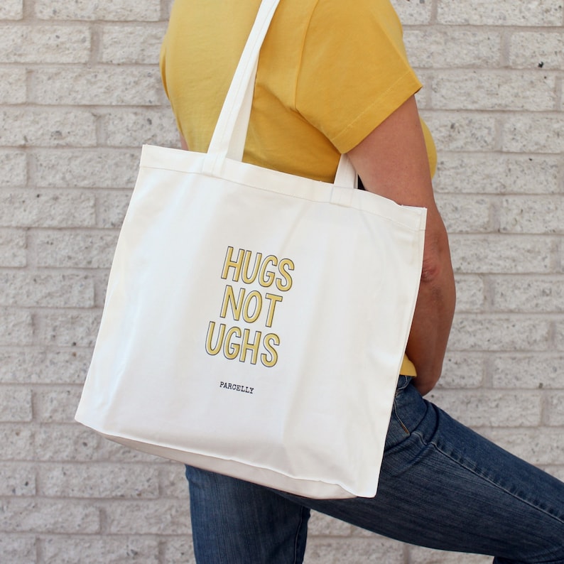May include: White canvas tote bag with yellow text that reads "Hugs Not Ughs" and the word "Parcelly" in smaller text below.