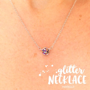 May include: A silver chain necklace with a small round pendant filled with colorful glitter. The pendant is about the size of a dime. The necklace is on a model's neck. The text "glitter necklace" is written in white letters on a white background below the image.