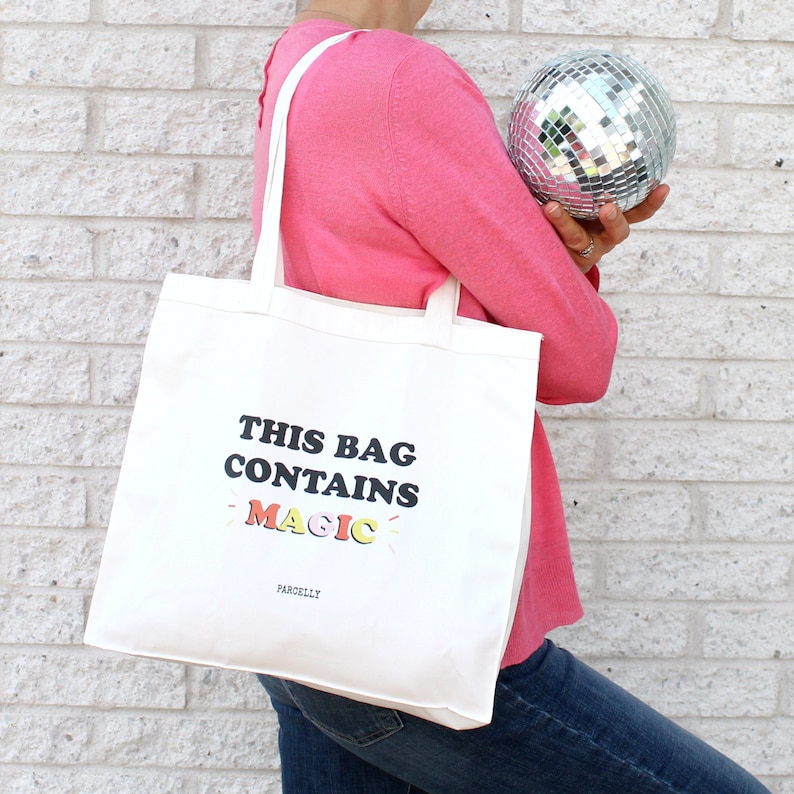 May include: A white canvas tote bag with the text "This bag contains magic" in black letters with rainbow accents. The text "Parcelly" is printed in black below the main text.