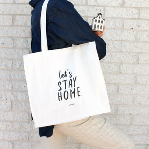 May include: White canvas tote bag with black text that reads "Let's Stay Home" and the word "Parcels" in smaller font below. The bag is being held by a person wearing a dark blue shirt.