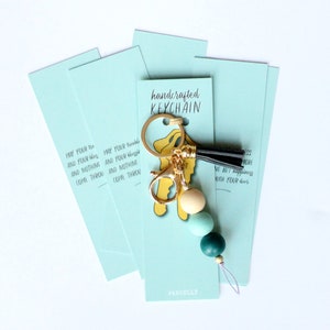 May include: A teal blue keychain with a gold key charm and a black tassel. The keychain has a quote that reads "May your troubles and your blessings come through and nothing come through". The keychain is handcrafted and comes with a teal blue card that says "handcrafted keychain" and "PAROELLY".