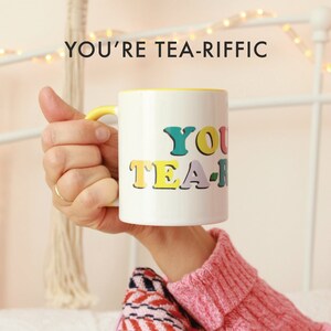 May include: White ceramic mug with a yellow rim and handle. The mug has the text "YOU'RE TEA-RIFFIC" in colorful letters.