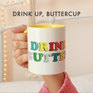 May include: White ceramic mug with a yellow rim. The mug has the text "DRINK UP, BUTTERCUP" printed on it in rainbow colors.