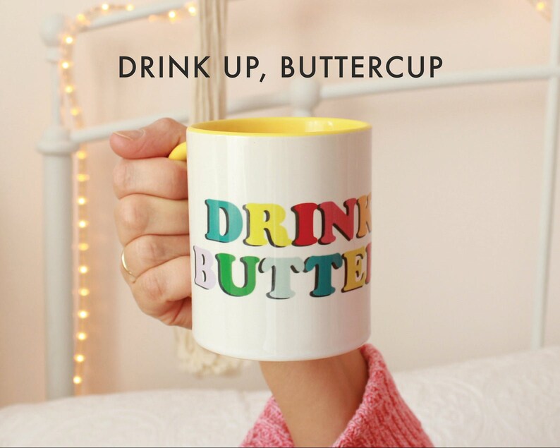 May include: White ceramic mug with a yellow rim. The mug has the text "DRINK UP, BUTTERCUP" printed on it in rainbow colors.