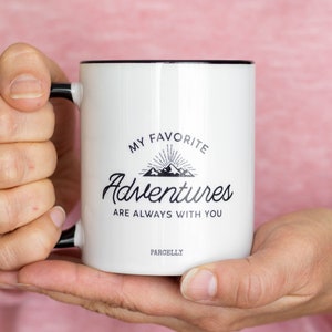 May include: White ceramic mug with a black rim and handle. The mug has a black graphic that says "My Favorite Adventures Are Always With You" with a mountain range and sunburst design. The text "ParcellY" is printed on the bottom of the mug.