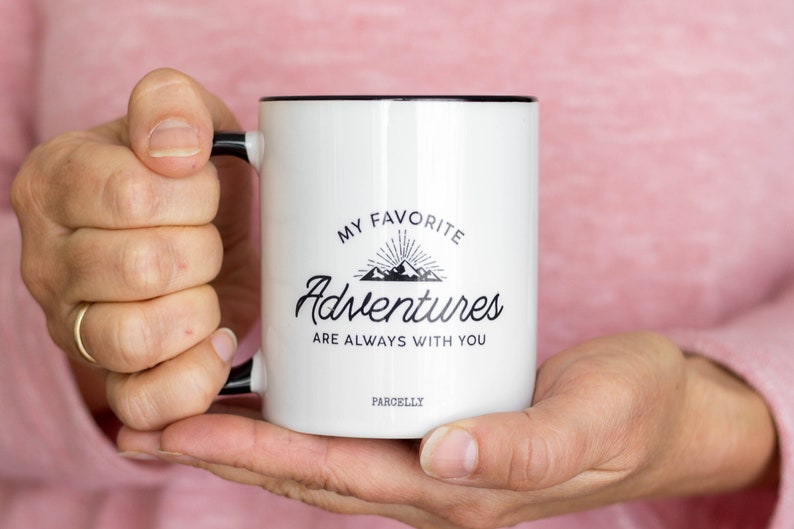May include: White ceramic mug with a black rim and handle. The mug has a black graphic that says "My Favorite Adventures Are Always With You" with a mountain range and sunburst design. The text "ParcellY" is printed on the bottom of the mug.