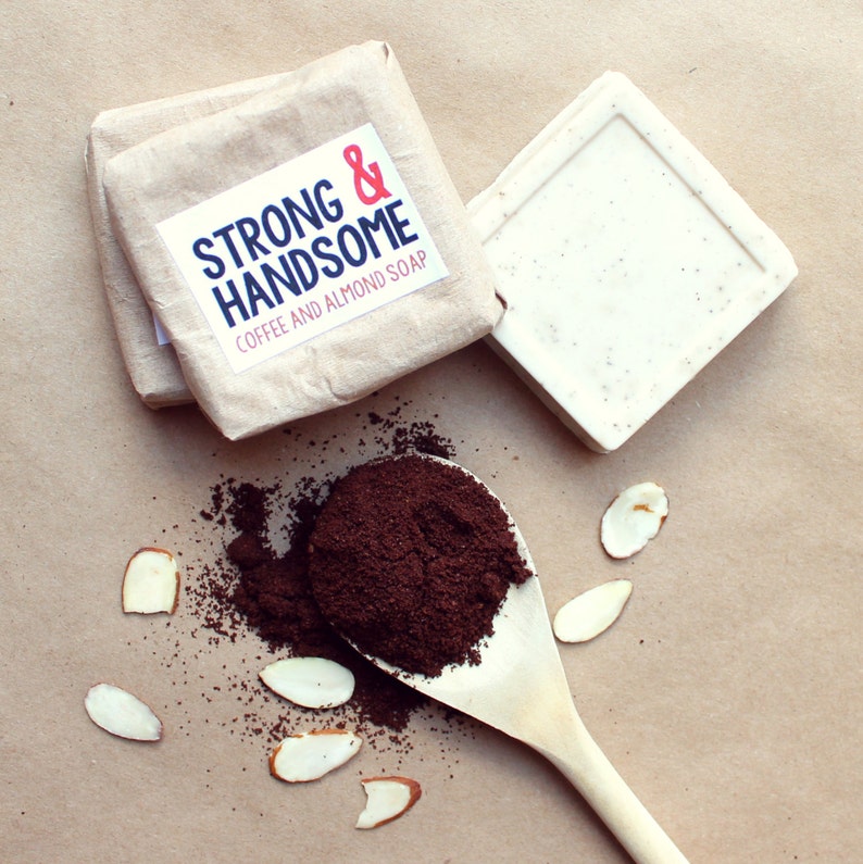 May include: Two bars of coffee and almond soap, one wrapped in brown paper with a label that reads "Strong & Handsome." A wooden spoon holds coffee grounds, with almond slices scattered around.