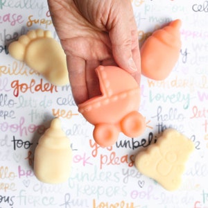 May include: A set of four baby-themed soap bars. The soaps are shaped like a baby bottle, a baby foot, a baby carriage, and a teddy bear. The soaps are all in pastel colors.
