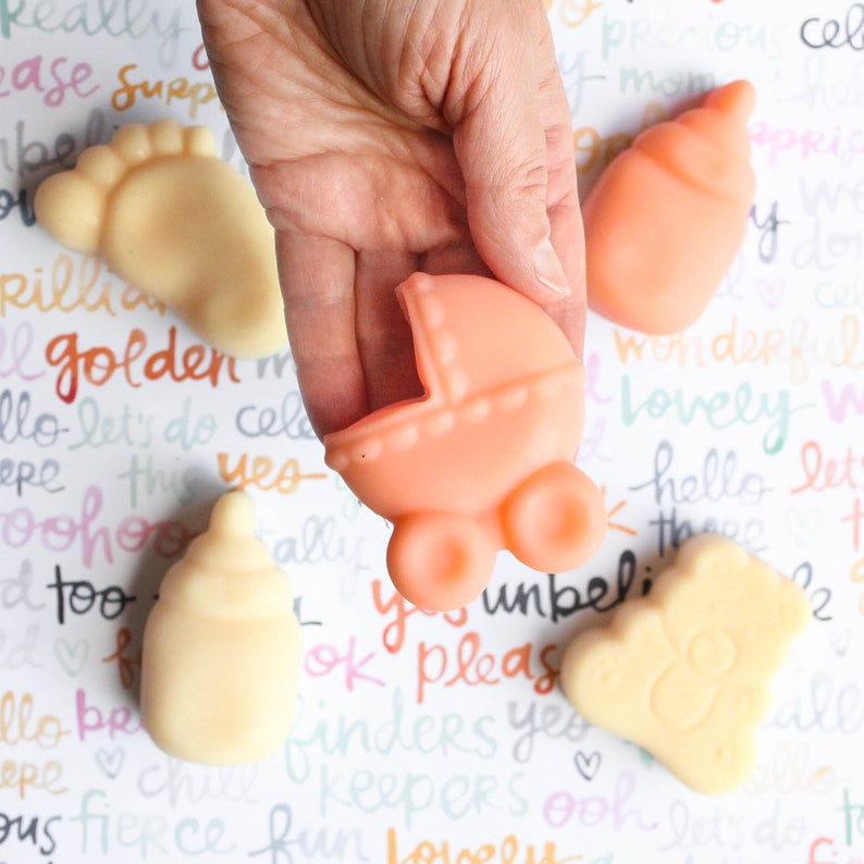 May include: A set of four baby-themed soap bars. The soaps are shaped like a baby bottle, a baby foot, a baby carriage, and a teddy bear. The soaps are all in pastel colors.