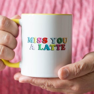 May include: White ceramic mug with a yellow handle and interior. The mug features the text "MISS YOU A LATTE" in colorful, playful lettering. The mug is held in two hands against a pink background.