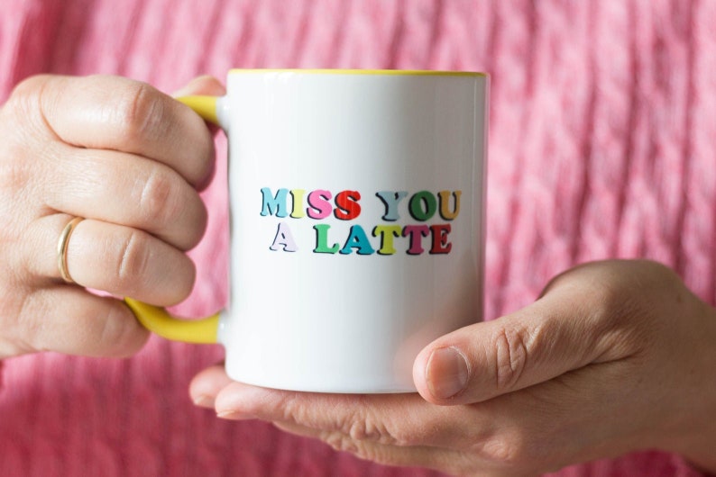 May include: White ceramic mug with a yellow handle and interior. The mug features the text "MISS YOU A LATTE" in colorful, playful lettering. The mug is held in two hands against a pink background.