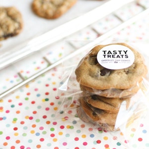 May include: A stack of four chocolate chip cookies in a clear plastic bag with a white circle sticker that says "Tasty Treats Chocolate Chip Cookies Made with Love" on a white background with colourful polka dots.