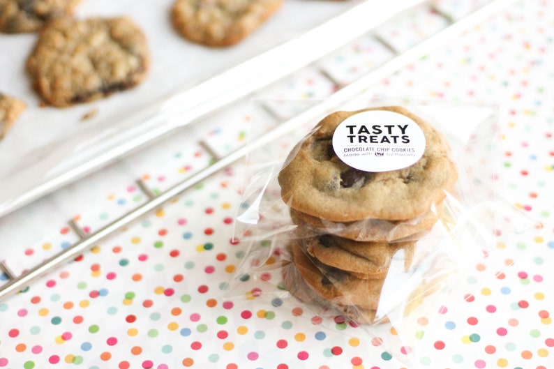 May include: A stack of four chocolate chip cookies in a clear plastic bag with a white circle sticker that says "Tasty Treats Chocolate Chip Cookies Made with Love" on a white background with colourful polka dots.