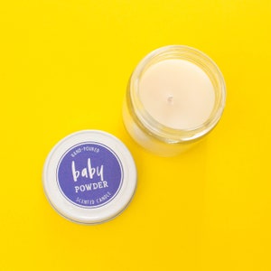 May include: A white candle jar with a purple label that reads "HAND-POURED Baby POWDER SCENTED CANDLE". The candle is in a clear glass jar and is sitting on a yellow surface.