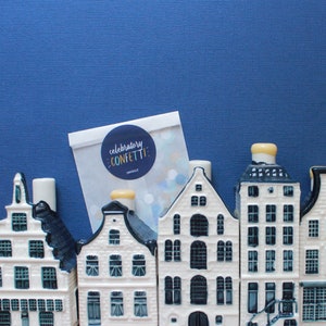 May include: A set of five white ceramic houses with blue trim and black windows. The houses are arranged in a row on a blue background. A white card with the text "celebratory confetti" is in the background.