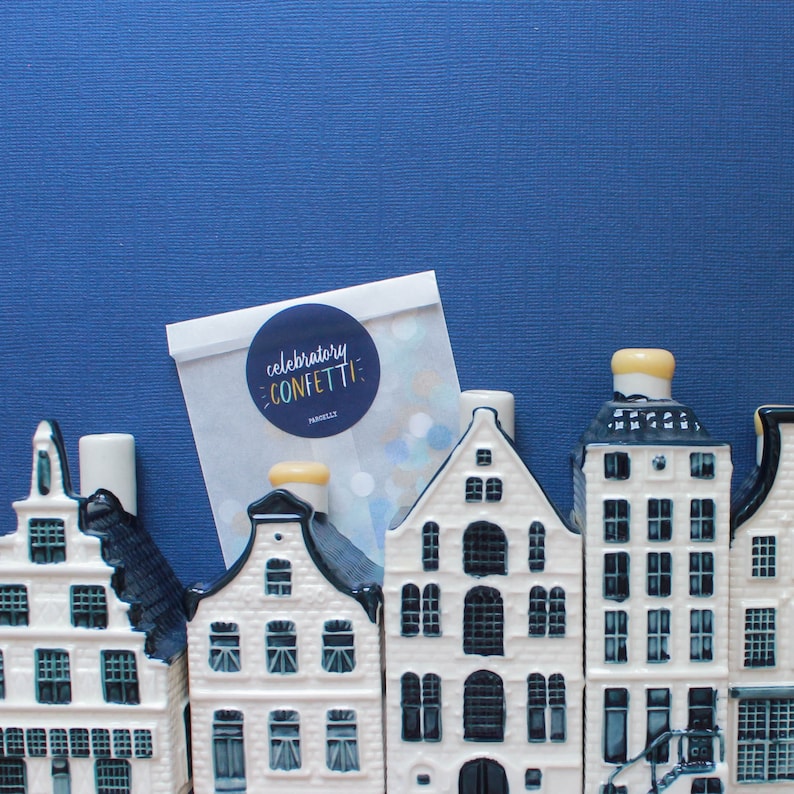 May include: A set of five white ceramic houses with blue trim and black windows. The houses are arranged in a row on a blue background. A white card with the text "celebratory confetti" is in the background.