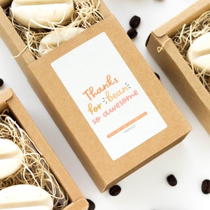 May include: A set of four small brown boxes with lids, each containing two white coffee bean-shaped soaps. The boxes are filled with shredded brown paper. One box has a sticker that says "Thanks for bean so awesome".
