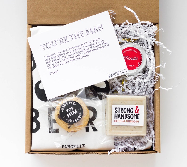 May include: A gift box containing a white t-shirt with black text, a cookie, a candle, and a bar of soap. The soap is labeled "Strong & Handsome Coffee and Almond Soap." A card reads "You're the Man."