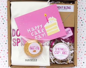 May include: A gift box with a birthday theme. It contains a white t-shirt with text, a pink card that says "Happy Cake Day!", a cookie, a necklace, and a candle. The candle is in a tin that says "Sprinkled Cupcake".