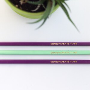 May include: Three pencils with purple and green painted bodies and gold lettering that reads "Grandparents to be".