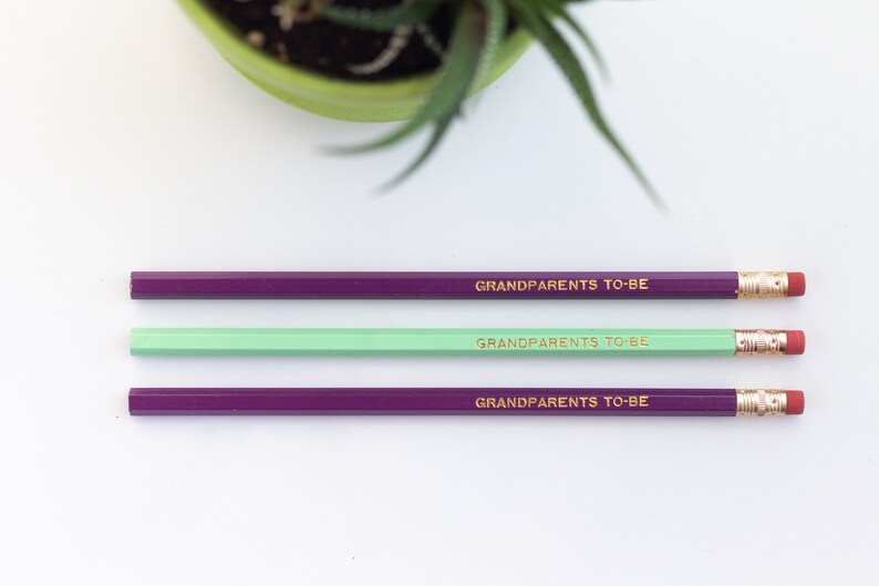 May include: Three pencils with purple and green painted bodies and gold lettering that reads "Grandparents to be".
