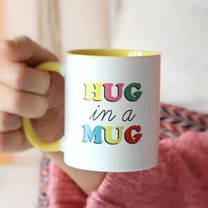May include: White ceramic mug with a yellow rim. The mug has the text "HUG in a MUG" written in colourful letters.