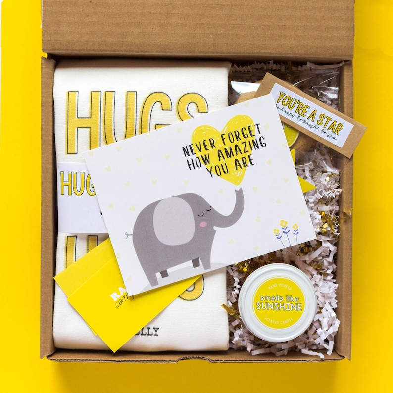 May include: A gift box filled with yellow and white themed items. The box contains a white t-shirt with the word "HUGS" printed in yellow, a card with an elephant holding a yellow heart that says "Never forget how amazing you are", a small jar of candle that says "smells like sunshine", and a small card that says "You're a star, be happy be bright be you".