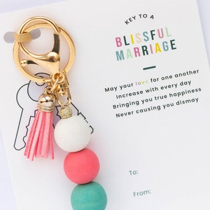 May include: Keychain with a pink tassel and four wooden beads in white, pink, teal, and white. The keychain is attached to a card that reads "Key to a Blissful Marriage" and includes a message about love and happiness.