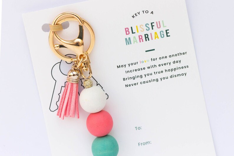 May include: Keychain with a pink tassel and four wooden beads in white, pink, teal, and white. The keychain is attached to a card that reads "Key to a Blissful Marriage" and includes a message about love and happiness.