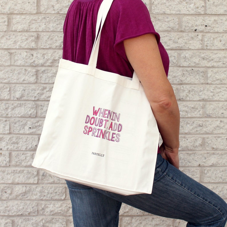 May include: White canvas tote bag with the text "When in doubt, add sprinkles" in pink and purple lettering. The text "Pardelly" is printed in smaller black lettering below the main text.