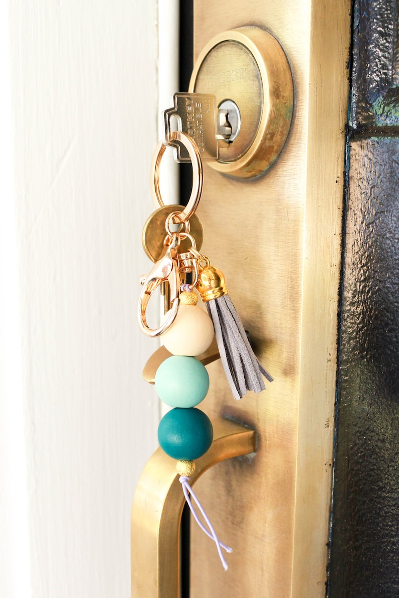 May include: A gold key chain with a gray tassel and three wooden beads in shades of teal and cream.