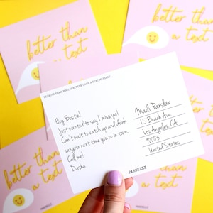 May include: A pink and yellow postcard with the text "better than a text" and a smiley face. The back of the postcard has a handwritten message that says "Hey Bestie! Just wanted to say I miss ya! Can't wait to catch up and drink sangria next time you're in town. Call me! Dasha" and an address for Madi Parslow.