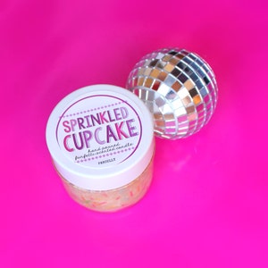 May include: A white jar with a pink and purple label that reads "SPRINKLED CUPCAKE hand poured confetti-scented candle PARCELLY". The jar is filled with a white substance with colorful sprinkles. A silver disco ball is in the background.
